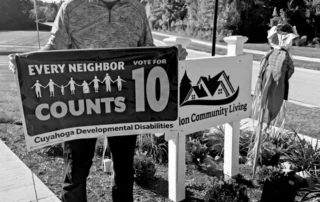 Ara Bagdasarian holds an Every Neighbor Counts Issue 10 sign beside the Solon Community Living sign
