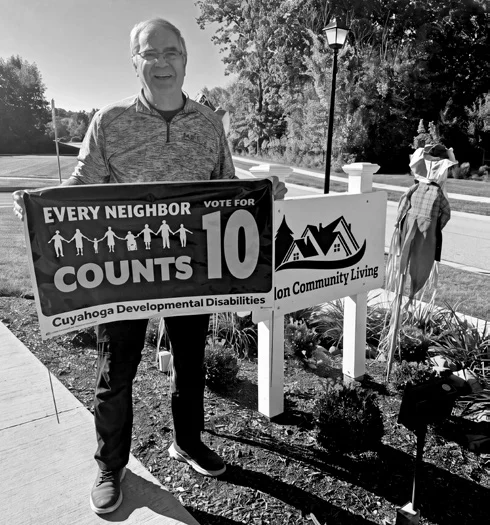Ara Bagdasarian holds an Every Neighbor Counts Issue 10 sign beside the Solon Community Living sign